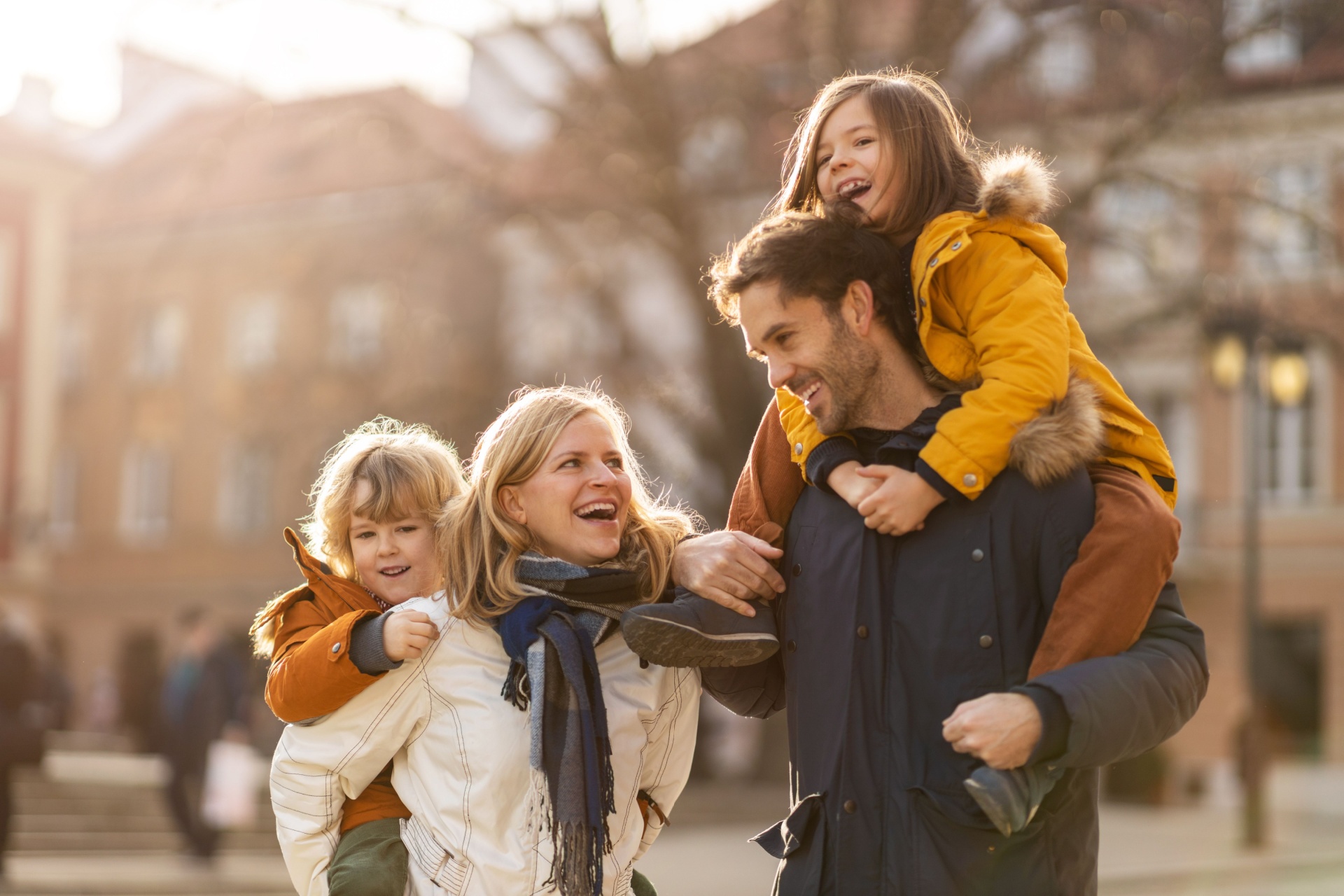A husband and wife both give piggybacks to their young children in the middle of a city.